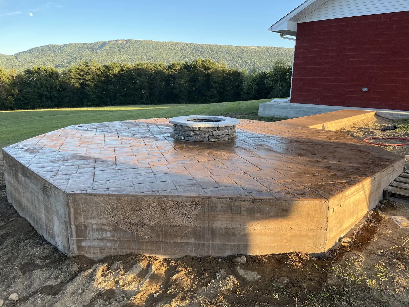 Raised stamped and stained concrete patio with a stone fire pit beside a red house